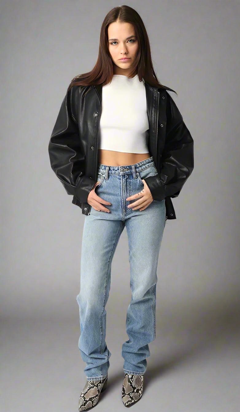 Woman wearing a black leather jacket, white crop top, and blue jeans on a neutral background